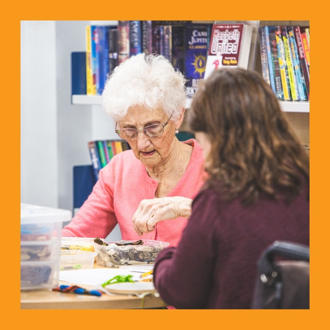 A photo of a woman with white hair and a woman with brown hair in a wheelchair, both working on a craft at a table in the library