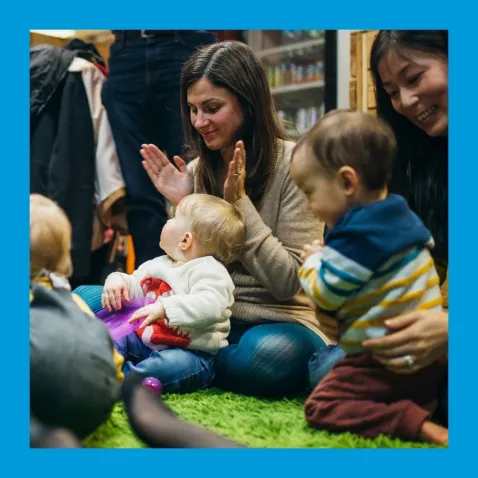 A photo of toddlers sitting on the floor in parents' laps, clapping and smiling