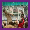 A storyteller sits in a chair in front of a group of children with raised hands while a dancer poses beside him
